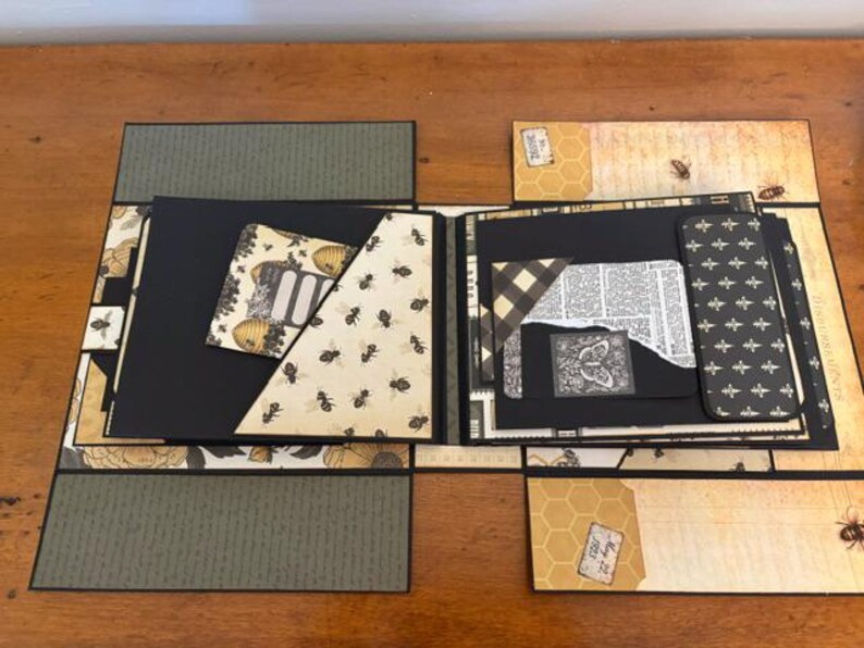 Pu&ograve; includere: Un album di ritagli artigianale con pagine nere, inserti di carta con motivi di api, disegni a nido d'ape ed elementi decorativi vintage. L'album aperto mostra varie tasche, texture di carta stratificate in tonalit&agrave; nero, crema e verde oliva.
