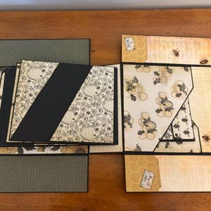 Pu&ograve; includere: Un album di ritagli a pi&ugrave; pannelli con tema api e alveari, caratterizzato da strati di cartoncino nero, carta color crema con illustrazioni di api e accenti verde oliva. Il design include tasche diagonali, sezioni pieghevoli ed etichette.