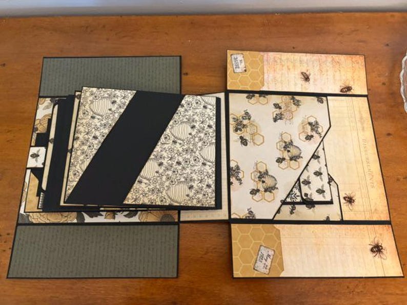Pu&ograve; includere: Un album di ritagli a pi&ugrave; pannelli con tema api e alveari, caratterizzato da strati di cartoncino nero, carta color crema con illustrazioni di api e accenti verde oliva. Il design include tasche diagonali, sezioni pieghevoli ed etichette.