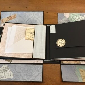 May include: A multi-panel scrapbook album featuring black cardstock pages and decorative paper accents in shades of light blue, cream, and tan. The album includes a white envelope tucked into a pocket and a circular sticker on a black flap.