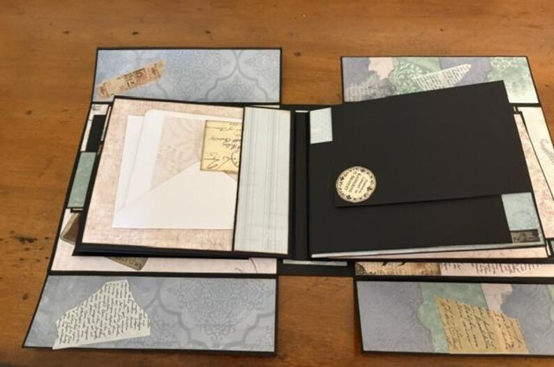 May include: A multi-panel scrapbook album featuring black cardstock pages and decorative paper accents in shades of light blue, cream, and tan. The album includes a white envelope tucked into a pocket and a circular sticker on a black flap.