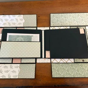 Op de afbeelding: Een handgemaakt plakboek met zwart kartonnen pagina's en meerdere uitklapbare flappen. Het papier heeft patronen in saliegroen, zachtroze en wit, waaronder botanische prints, geometrische rasters en damastontwerpen met zwarte randen.