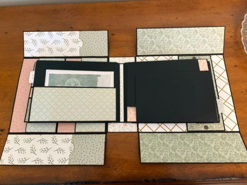 Op de afbeelding: Een handgemaakt plakboek met zwart kartonnen pagina's en meerdere uitklapbare flappen. Het papier heeft patronen in saliegroen, zachtroze en wit, waaronder botanische prints, geometrische rasters en damastontwerpen met zwarte randen.