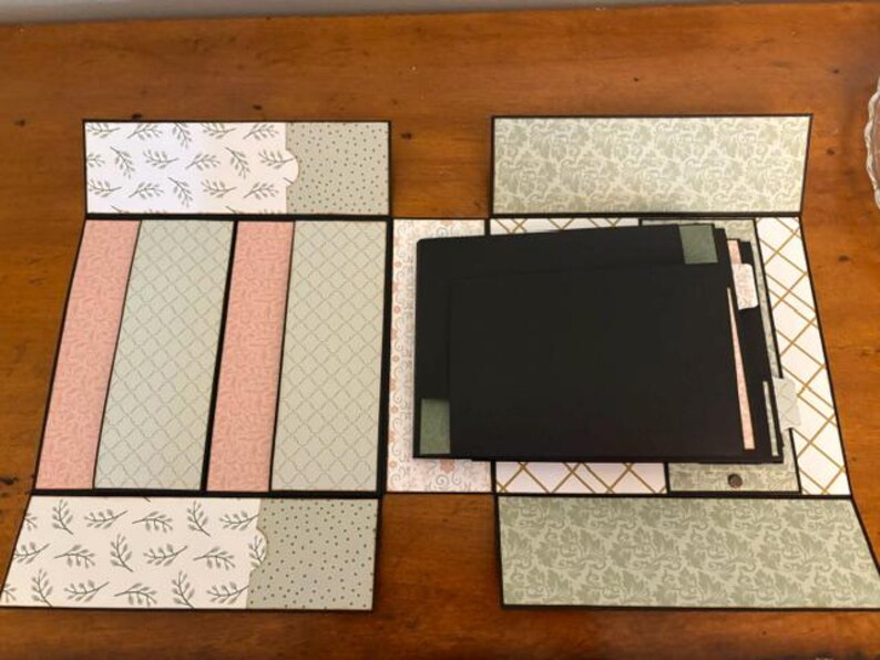 Op de afbeelding: Een meerdelig plakboekalbum met diverse patronen in saliegroen, zachtroze en wit. Het open album toont verschillende zwarte kartonnen pagina's en vakjes, voorzien van decoratieve blad-, ruit- en bloemenprints.