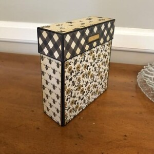 Pu&ograve; includere: Una scatola rettangolare con coperchio a motivo vichy bianco e nero. I lati sono decorati con carta color crema stampata con api nere e un motivo floreale di fiori dorati e neri. Un piccolo porta etichette in metallo dorato si trova sul coperchio.