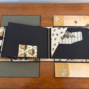Pu&ograve; includere: Un album di ritagli artigianale con pagine nere e accenti di carta decorativa con motivi di api, favi e note musicali. L'album aperto mostra molteplici pannelli pieghevoli, tasche ed elementi in cartoncino stratificato con illustrazioni di api in stile vintage ed etichette di testo su una struttura multistrato.