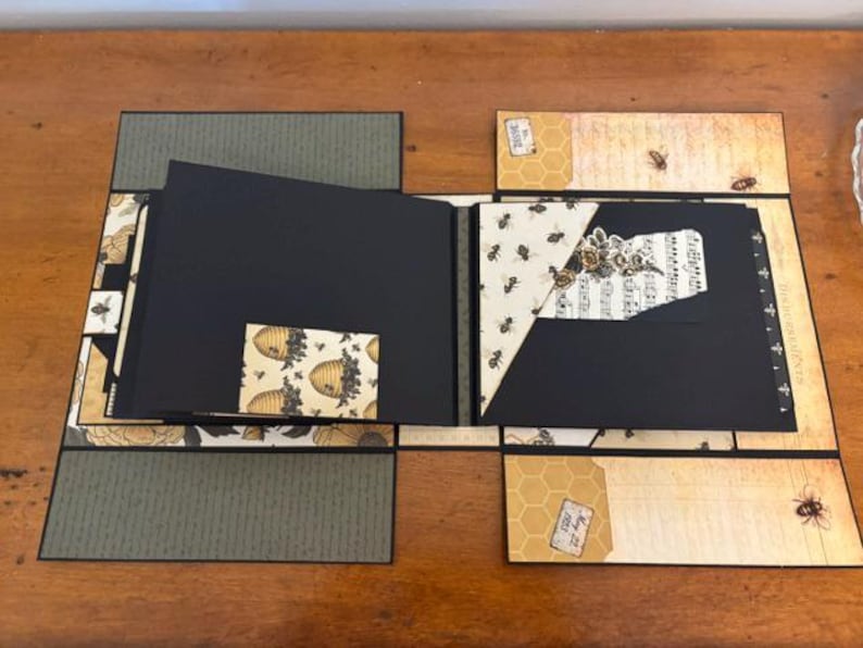 Pu&ograve; includere: Un album di ritagli artigianale con pagine nere e accenti di carta decorativa con motivi di api, favi e note musicali. L'album aperto mostra molteplici pannelli pieghevoli, tasche ed elementi in cartoncino stratificato con illustrazioni di api in stile vintage ed etichette di testo su una struttura multistrato.