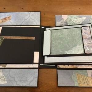 May include: A multi-layered scrapbook album featuring black cardstock pages and decorative paper accents in shades of blue, green, and cream. The album includes fold-out panels, pockets, and vintage-style printed ephemera with script text.