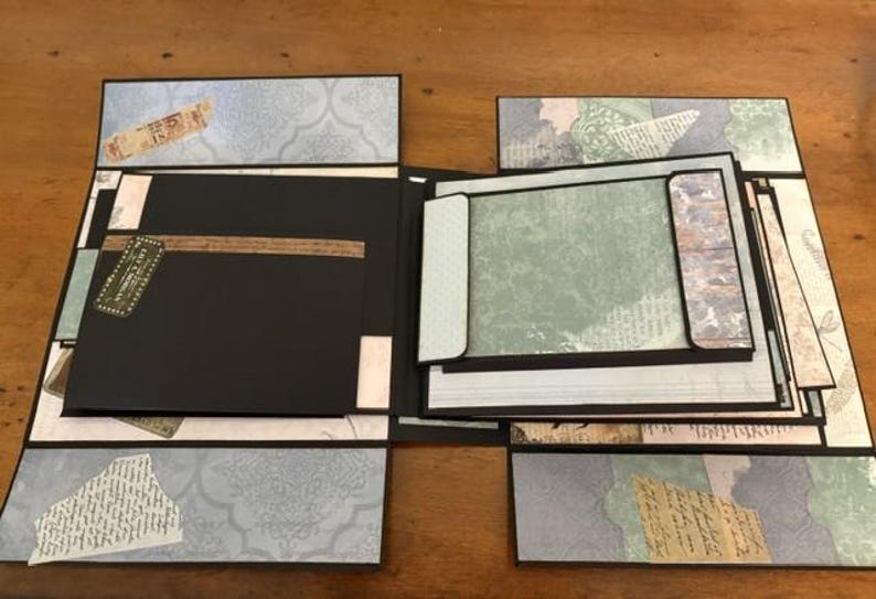 May include: A multi-layered scrapbook album featuring black cardstock pages and decorative paper accents in shades of blue, green, and cream. The album includes fold-out panels, pockets, and vintage-style printed ephemera with script text.