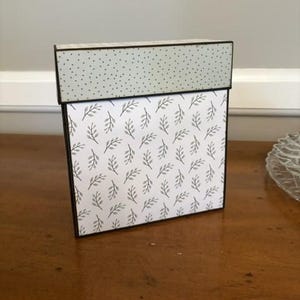 Op de afbeelding: Een vierkante doos met een deksel met een lichtgroene achtergrond met kleine donkere stippen. Het hoofdgedeelte van de doos is wit met een herhalend patroon van kleine grijze bladeren. De randen van de doos en het deksel zijn donkerbruin.