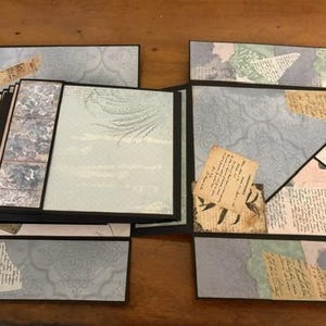 May include: A multi-layered scrapbook album featuring a black frame and various panels decorated with light blue, cream, and sage green patterned paper. The design includes collage elements, vintage-style script text, and botanical illustrations.