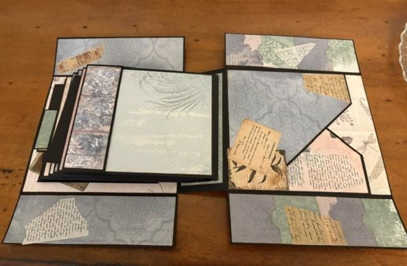 May include: A multi-layered scrapbook album featuring a black frame and various panels decorated with light blue, cream, and sage green patterned paper. The design includes collage elements, vintage-style script text, and botanical illustrations.