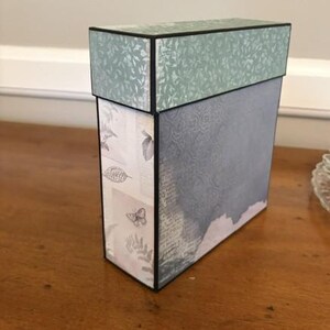 May include: A rectangular storage box with a lid, featuring a sage green leaf pattern on the top and a blue-grey textured surface on the side. The front panel displays a light background with printed botanical illustrations and butterfly motifs.