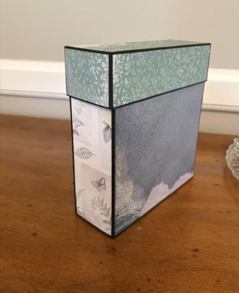 May include: A rectangular storage box with a lid, featuring a sage green leaf pattern on the top and a blue-grey textured surface on the side. The front panel displays a light background with printed botanical illustrations and butterfly motifs.