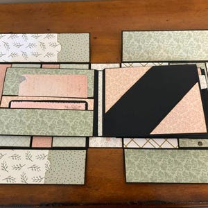Op de afbeelding: Een handgemaakt plakboekalbum met meerdere uitklapbare panelen en vakjes. Het ontwerp bevat zwart karton met daarop bedrukt papier in tinten saliegroen, zachtroze en wit, voorzien van bloemmotieven, damastpatronen en geometrische vormen.
