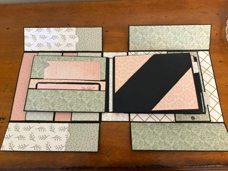 Op de afbeelding: Een handgemaakt plakboekalbum met meerdere uitklapbare panelen en vakjes. Het ontwerp bevat zwart karton met daarop bedrukt papier in tinten saliegroen, zachtroze en wit, voorzien van bloemmotieven, damastpatronen en geometrische vormen.