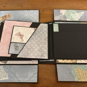 May include: A multi-layered scrapbook album featuring black cardstock pages with decorative paper inserts in shades of blue, pink, and green. The album includes various pockets, fold-out sections, and collage-style paper accents with butterfly motifs.