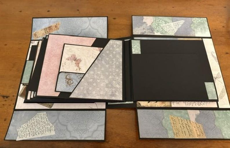May include: A multi-layered scrapbook album featuring black cardstock pages with decorative paper inserts in shades of blue, pink, and green. The album includes various pockets, fold-out sections, and collage-style paper accents with butterfly motifs.