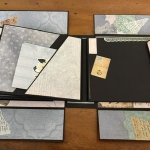 May include: A multi-panel scrapbook with black cardstock pages and decorative paper inserts in shades of blue, green, and cream. The layout features various pockets, flaps, and layered paper elements with printed patterns, script text, and floral motifs.