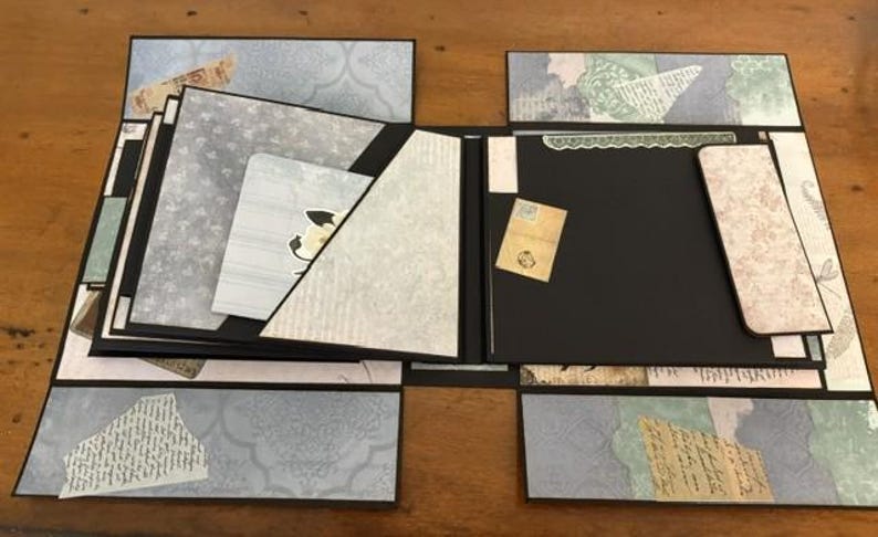 May include: A multi-panel scrapbook with black cardstock pages and decorative paper inserts in shades of blue, green, and cream. The layout features various pockets, flaps, and layered paper elements with printed patterns, script text, and floral motifs.