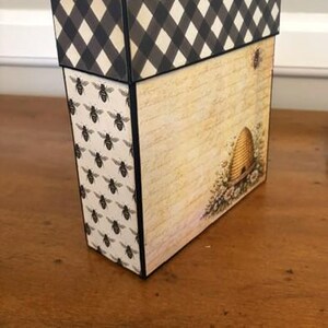 Pu&ograve; includere: Una scatola decorativa rettangolare con un coperchio a quadretti bianchi e neri. Il pannello laterale presenta un motivo ripetuto di piccole api nere su sfondo crema, mentre il pannello frontale mostra uno sfondo con scrittura vintage.