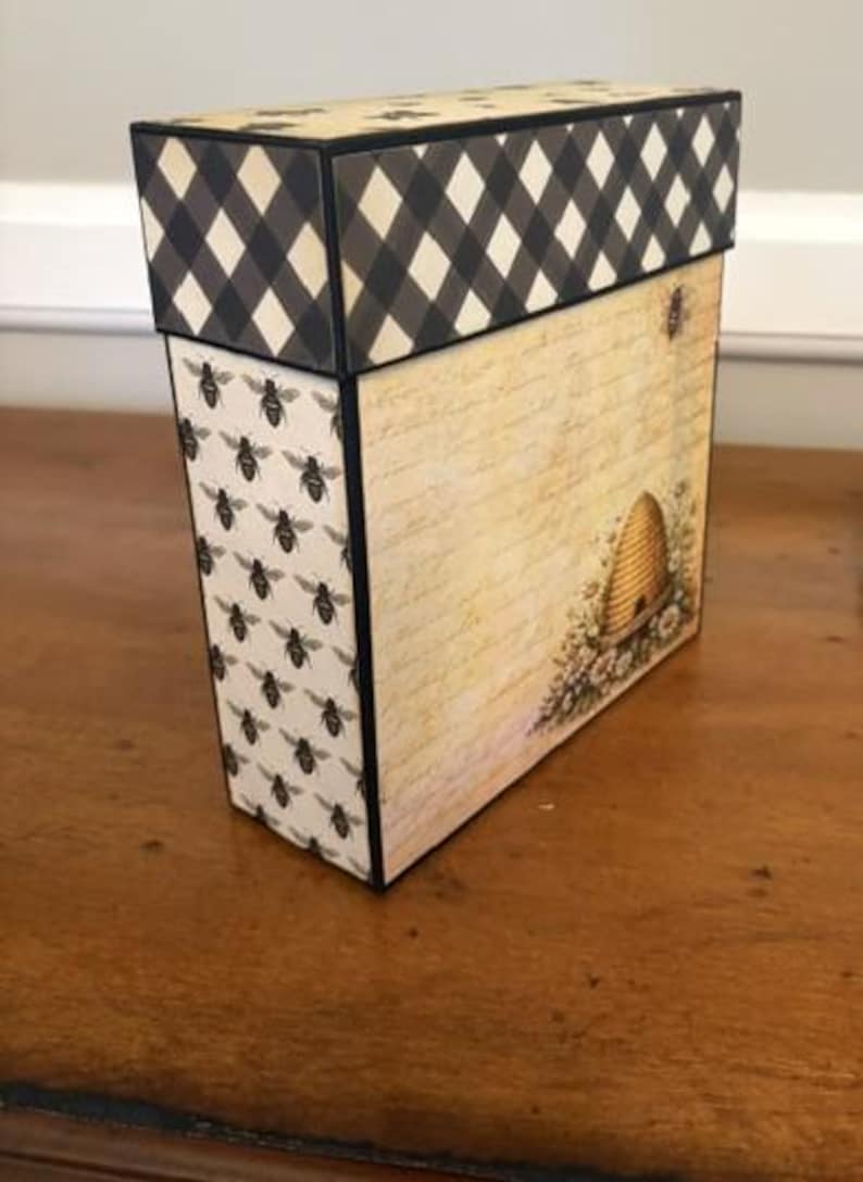 Pu&ograve; includere: Una scatola decorativa rettangolare con un coperchio a quadretti bianchi e neri. Il pannello laterale presenta un motivo ripetuto di piccole api nere su sfondo crema, mentre il pannello frontale mostra uno sfondo con scrittura vintage.