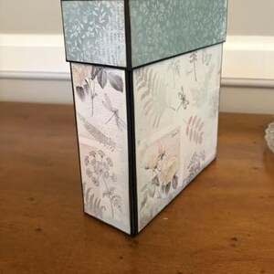 May include: A rectangular storage box with a lid, featuring a light green and cream colour palette. The exterior is decorated with botanical illustrations of ferns, flowers, and dragonflies, accented by black edges and a textured floral pattern on the lid.