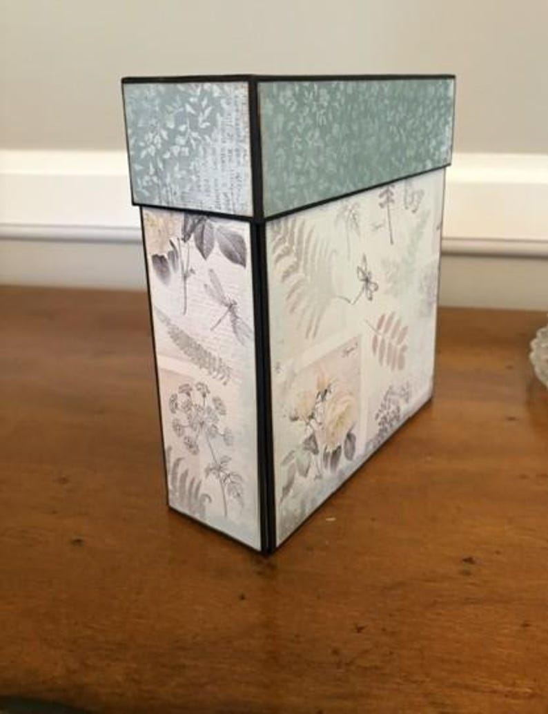 May include: A rectangular storage box with a lid, featuring a light green and cream colour palette. The exterior is decorated with botanical illustrations of ferns, flowers, and dragonflies, accented by black edges and a textured floral pattern on the lid.