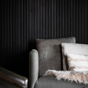 May include: A gray armchair with a fluffy throw blanket and several pillows. The armchair is in front of a black wood wall with vertical panels.