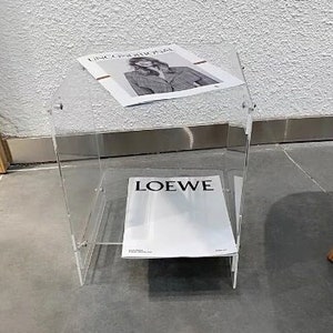 Puede incluir: Una mesa auxiliar transparente de acrílico con una revista encima y otra en el estante inferior. La revista superior tiene la palabra "UNCONDITIONAL" impresa. La revista inferior tiene la palabra "LOEWE".