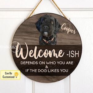 May include: A round wooden sign with a faux wood grain finish. The sign features a black and white photo of a dog with the text "Cooper" in pink script. The sign also includes the text "Welcome-ISH DEPENDS ON WHO YOU ARE & IF THE DOG LIKES YOU".
