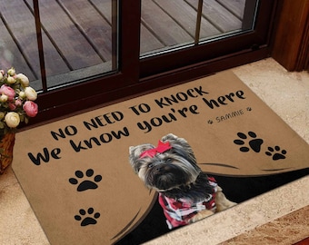 以下が含まれることがあります： 「No need to knock we know you're here」という文字と、赤いリボンをつけた黒と茶色の小さな犬が描かれた茶色の玄関マット。犬の名前は「Sammie」で、玄関マットには足跡があります。
