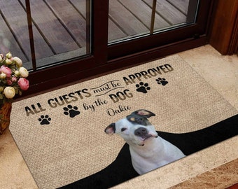 以下が含まれることがあります： 「ALL GUESTS must be APPROVED by the DOG」の文字と足跡がプリントされたベージュのドアマット。オークリーという名前の白黒の犬がマットに描かれています。マットの下部には黒い縁取りがあります。