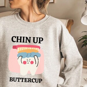 以下が含まれることがあります： 「Chin Up Buttercup」というテキストと、漫画の歯のグラフィックが描かれた灰色のスウェットシャツです。