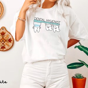 以下が含まれることがあります： 歯ブラシを持った3つの漫画の歯のグラフィックが描かれた白いTシャツ。歯の上に「歯科衛生士」という文字があります。