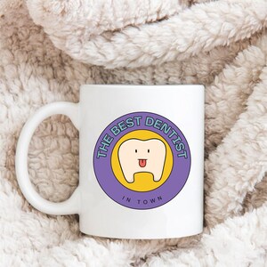 May include: White ceramic mug with a purple circle design featuring a cartoon tooth with a tongue sticking out. The text "THE BEST DENTIST IN TOWN" is written around the tooth.