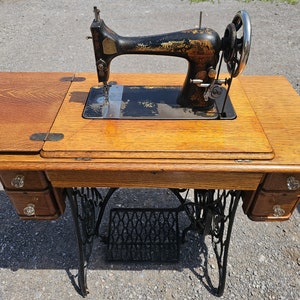 Op de afbeelding: Antiek zwarte naaimachine met een houten tafel en laden. De machine heeft een zwarte metalen basis en een zwarte metalen arm met een naald en draad.
