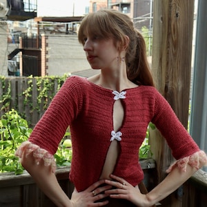 May include: A red crocheted cardigan with white crocheted flower accents. The cardigan has a deep V-neckline and is open in the front. The model is wearing a brown skirt.