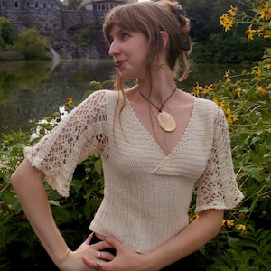 May include: A cream-coloured crochet top featuring a V-neckline, a fitted bodice, and flared, intricate lace-patterned sleeves. The garment is paired with a black cord necklace featuring a large, smooth, oval-shaped pendant.