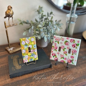 Puede incluir: Dos tapas de interruptor de luz pintadas con diseños florales. Una tapa presenta un fondo verde con flores rosas, amarillas y moradas. La otra tapa tiene un fondo blanco con rosas rosas. Ambas tapas están en soportes decorativos.