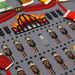 May include: A cross-stitch design on grey fabric depicting a burning building, skeleton figures, and decorative elements. The design uses red, black, orange, and white thread. Spools of thread in various colours surround the cross-stitch.