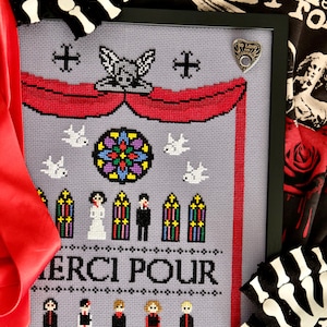 May include: Framed cross-stitch art with a gray background, featuring a red banner, stained glass, and the text "MERCI POUR." The design includes figures, doves, and a decorative angel. A pin with the text "SO LONG" is on the frame.