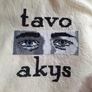Könnte beinhalten: Cremefarbener Stoff mit schwarzem Kreuzstich-Text und einem Augenbild. Der Text lautet "tavo akys" über und unter einem rechteckigen Bild von zwei Augen mit blauen Iris und Grautönen.