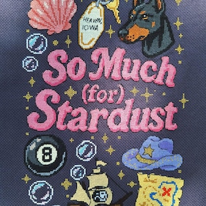 Pode incluir: Obra de arte em ponto cruz sobre um fundo azul escuro com uma concha rosa, uma etiqueta de chave que diz "Heaven, Iowa", uma cabeça de Doberman e a frase "So Much (for) Stardust". Outros elementos incluem uma bola 8, um navio e um mapa do tesouro.