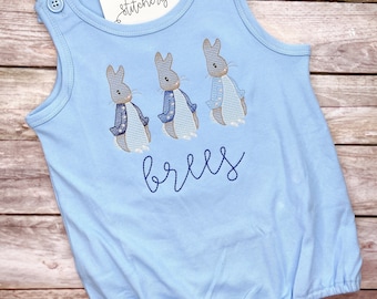 May include: Light blue baby onesie with three embroidered Peter Rabbit characters and the name "brees" embroidered below. The onesie has a sleeveless design and poppers at the bottom.