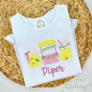 Könnte beinhalten: Weißes T-Shirt mit einem rosa Limonadenstand, einer Kanne und einem Glas Limonade, die darauf gestickt sind. Der Name "Piper" ist in rosa unter dem Limonadenstand gestickt.