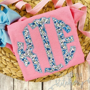 May include: Pink t-shirt with a large monogram in a floral print. The monogram is in a circle and the letters are "KF". The shirt has the text "The Little Stitchery" on the bottom.