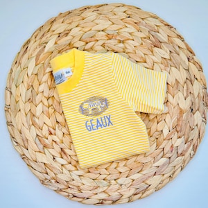 May include: A yellow and white striped baby shirt with the word "GEAUX" in blue. The shirt has a small graphic above the text and is folded on a woven, circular mat.