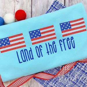 Könnte beinhalten: Ein hellblaues T-Shirt mit drei gestickten amerikanischen Flaggen und dem Text "Land of the Free" in blauer Farbe.