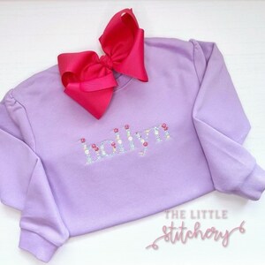 May include: A lavender sweatshirt with the name "Holly" spelled out in colorful embroidered flowers. A pink bow is on top of the sweatshirt.